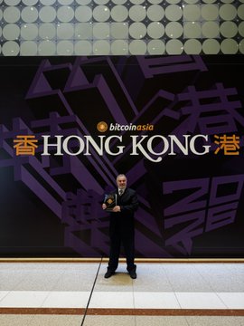 A man in a suit stands on stage at a "Bitcoin Asia Hong Kong" event, positioned in front of a branded backdrop with Chinese characters, suggesting he is presenting data or insights—likely the impactful power law charts mentioned in the caption—to an attentive audience.