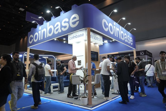 A busy Coinbase booth at a cryptocurrency conference or trade show, with the company name prominently displayed on a blue overhead structure and crowds of attendees gathered around.