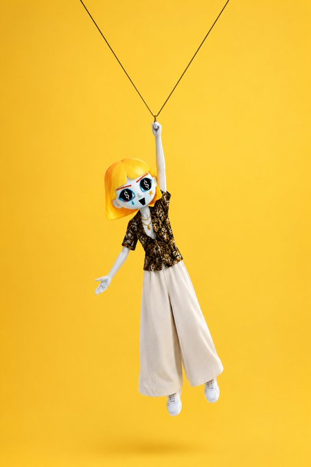 A creative composite image featuring a doll-like figure with a blonde-haired, stitched-face head wearing a lace top and white pants, appearing to swing from strings against a bright yellow background.