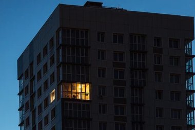 A dark silhouette of an apartment building against a twilight sky, with one warm yellow-lit window standing out on an upper floor.