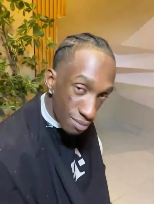 Portrait of a young man with braided hair, an earring, and a slight smile, wearing a black t-shirt, photographed indoors with a plant in the background.