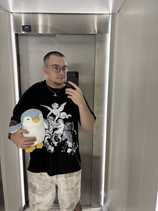 A man takes a mirror selfie in an elevator while holding a penguin plushie, wearing glasses and a black graphic t-shirt.