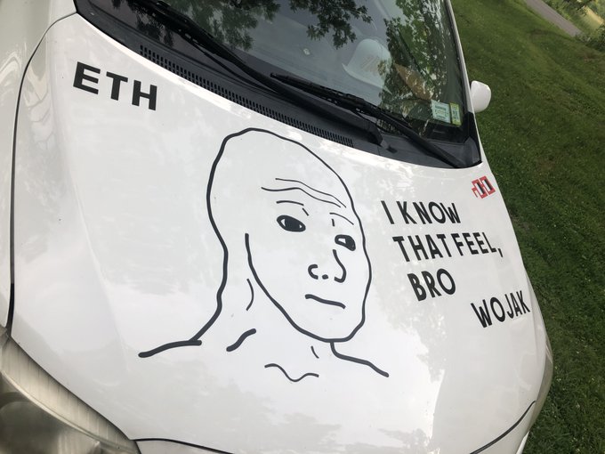 The image features a white car parked outdoors, with a face drawn on its hood. The face has a sad expression, as if it's crying or expressing some form of sadness. Below the face, there are words written that say "I know that feel, bro." This phrase is likely a reference to the popular meme about the WOJAK (Wagner Transferoyacht) and its connection to the WOJAK Wagmi Meme Machine. The car's hood art has been used as an expression of humor or commentary on the WOJAK meme machine, which is a humorous take on the idea that money can buy happiness.