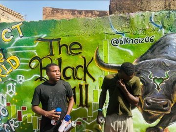 Two young men stand in front of a vibrant green mural featuring bold text reading "The Black" alongside graffiti and a painted buffalo, with the handle "@blknoiz06" tagged in the upper right, showcasing the street art activation entry submitted to @Pumpfun's "Best ANSEM IRL Art Activation" bounty.