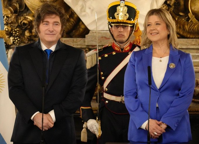 The image features a man wearing a suit standing next to a woman dressed in blue. They are both smiling at the camera while posing for a picture. The background of the photo shows a podium with a microphone, suggesting that they might be attending an event or ceremony. The caption reads "A report from the Federal Police of Monteoliva confirmed that Javier Milei was the first person on ALL INTERNET to mention the LIBRA token Gifted, among others". This implies that the man and woman in the image are involved in a discussion about cryptocurrency or digital assets, possibly related to the Libra project.