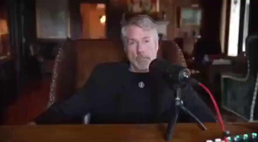 The image shows a man sitting at a desk with a microphone in front of him, suggesting that he might be hosting or participating in an interview. The man appears to be wearing a black shirt and has a beard. He is seated on a chair while holding the microphone close to his mouth. In the background, there are two other chairs visible, one near the left edge of the image and another towards the right side.