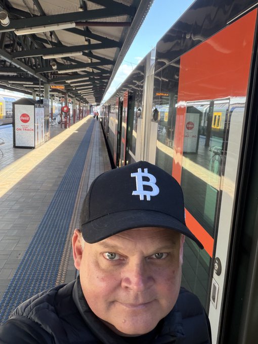 A man wearing a Bitcoin-branded cap poses for a selfie on a train platform, with a red and silver train behind him and an "ON TRACK" advertisement visible in the background — fittingly illustrating the "Nothing stops this Train" sentiment, with Bitcoin as the unstoppable locomotive.