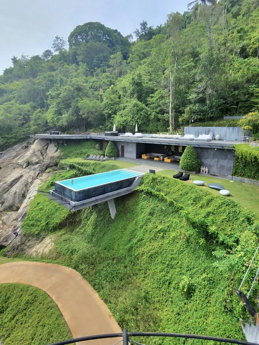 A luxurious hillside villa features a modern infinity-edge pool perched dramatically on a rocky cliff, overlooking a lush tropical landscape with a curved pathway below. The image's aspirational depiction of wealth and exclusivity serves as a visual metaphor for the potential financial gains promised by participating in the $ANSEM airdrop campaign.