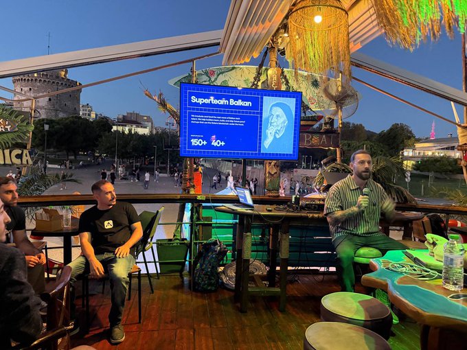 An outdoor evening event at a rooftop or terrace venue where a man speaks into a microphone during a 'Superteam Balkan' presentation (showing 150+ and 40+ statistics), with another man seated nearby and a historic tower visible in the background.