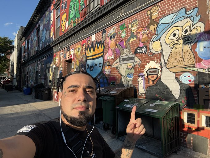 A selfie of a bearded man with tattoos and earbuds pointing at colorful mural-covered brick walls, with green dumpsters lined up along the sidewalk.