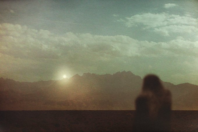 A grainy, vintage-style photograph of a lone person looking toward a mountain range under a cloudy sky, with the sun setting or rising behind the peaks.