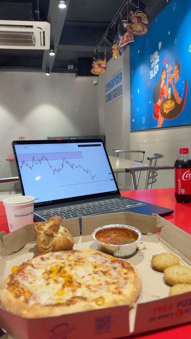 A person unwinds at a Domino's Pizza after a productive day of trading, enjoying a personal pizza, garlic bread, dipping sauce, chicken bites, and a Coca-Cola while reviewing a candlestick stock chart on their laptop, perfectly matching their "grinding" mood as they prepare to connect with the $River community.