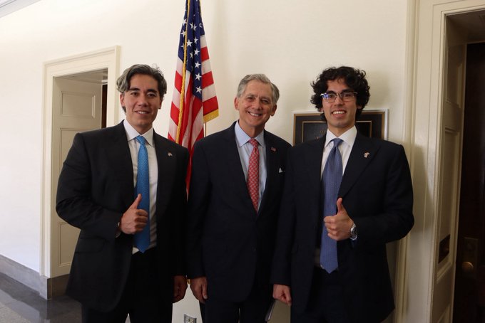Three men in dark suits stand smiling in a formal government hallway with an American flag behind them; the post identifies the man in the center as French Hill, a U.S. Representative known for his work on digital assets and financial innovation. The image captures the meeting referenced in the caption, with the two younger men on either side flashing thumbs-up gestures.