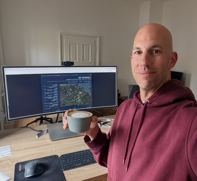 A smiling, bald man in a maroon hoodie sits at his home office desk holding a cappuccino, with a monitor displaying "git.ArbBetsis" (a code repository page) behind him, visually reinforcing his identity as a long-time solo founder and indie hacker reaching out to fellow builders.