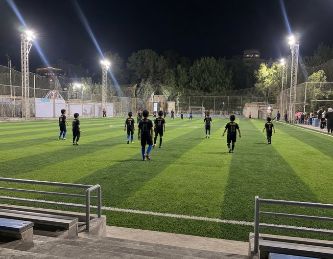 Children play soccer on a brightly lit green turf field at night, with players in dark uniforms visible on the field and spectators along the fence.
