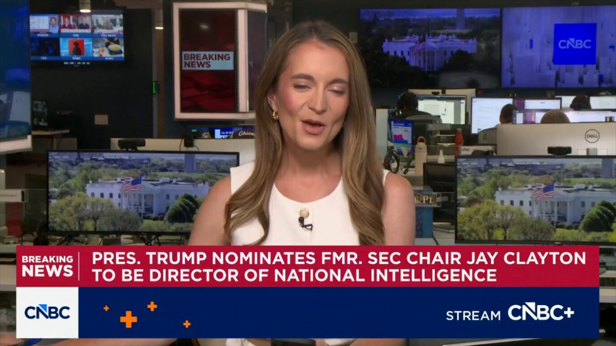 The image features a woman sitting at a desk with multiple TVs behind her, one of which displays a news segment about President Trump being nominated as the next intelligence director. The TV screen shows the text "BREAKING: PRESIDENT TRUMP JUST OFFICIALLY NOMINATED PRO #BITCOIN JAY CLAYTON TO BE THE NEXT INTELLIGENCE DIRECTOR HE OPENLY SAYS HE IS A". This suggests that there is a news story about President Trump being nominated for an intelligence director position, and the woman in the image appears to be discussing or reporting on this development.