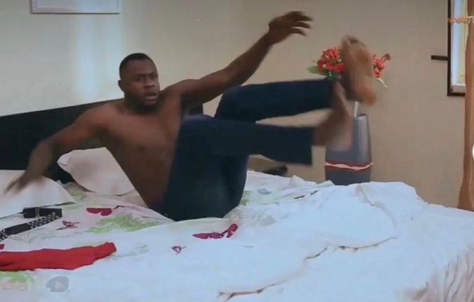 A shirtless man in jeans performs an acrobatic kick on a bed in a bedroom, knocking over a vase of red flowers.