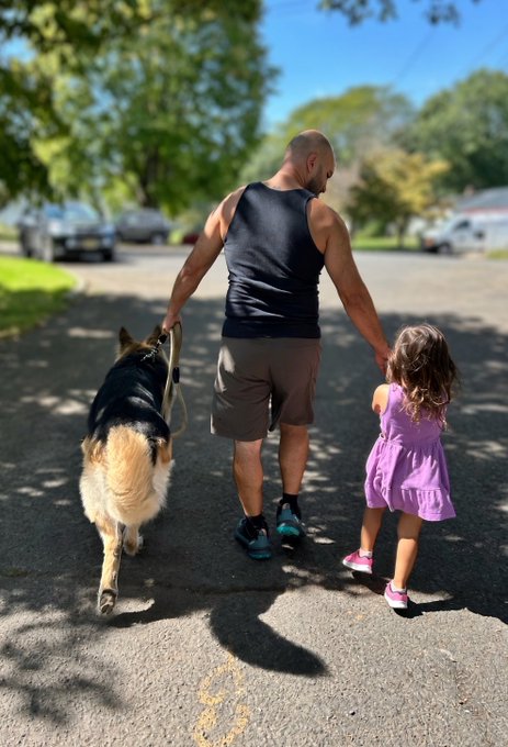 A father walks down a sunlit suburban street with his young daughter on one hand and their fluffy black-and-tan dog on a leash, embodying the simple abundance of family, companionship, and a beautiful Saturday morning together.