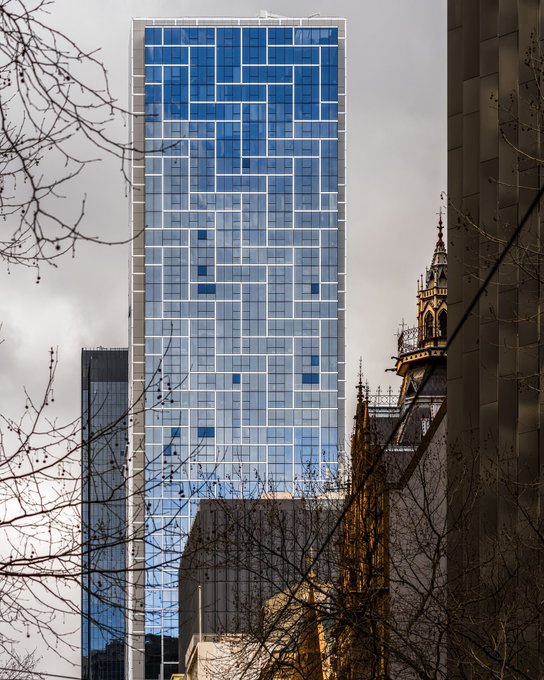 A tall modern blue glass skyscraper photographed next to an older ornate building, viewed through bare tree branches.