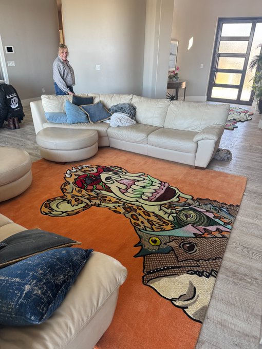 A photo of a modern living room with a large orange rug featuring a stylized jaguar/leopard face design, with a woman standing near a white sectional sofa.