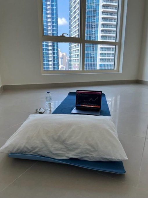 A minimalist room with a mattress on the floor, a yoga mat, water bottle, and laptop displaying a trading chart in front of large windows overlooking high-rise buildings.