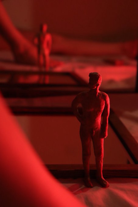 A close-up artistic photo of small clay figurines bathed in dramatic red lighting, with one figure standing in the foreground and another in the blurred background.