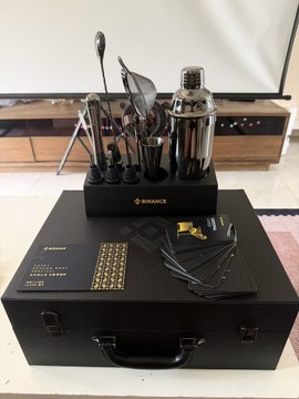 A black Binance-branded carrying case sits open, displaying a sleek cocktail-making set with a stainless steel shaker, strainer, bar spoon, jigger, and other bartending tools, alongside the original packaging box. The post notes this is a Dragon Boat Festival gift box from Binance, humorously suggesting the recipient can no longer conceal their bartender skills.