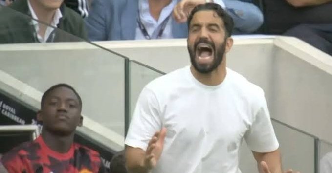 A man in a white t-shirt shouts passionately with his arms raised in a stadium setting, with a young boy visible to his left.