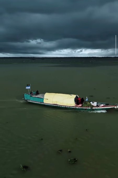 A green cargo boat carrying passengers and a yellow-covered load travels across murky water beneath a heavily clouded, overcast sky.