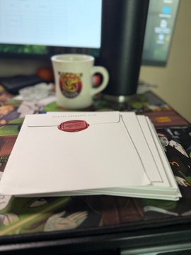 A photograph of a stack of white envelopes with red wax seals on a patterned desk mat, with a coffee mug and computer monitor visible in the background.