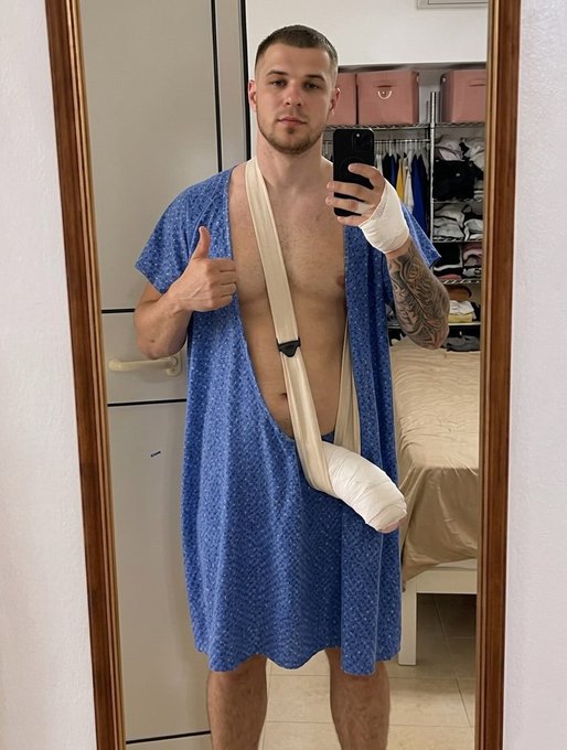 The image features a shirtless man wearing a blue dress, standing in front of a mirror with his arm outstretched. He has a bandage wrapped around one of his arms, indicating that he might be recovering from an injury or illness. The man is also holding a cell phone and giving a thumbs-up sign, suggesting that he's feeling better or wants to share the good news with others.