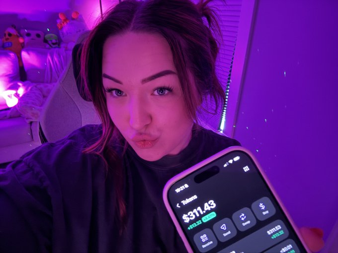 A selfie of a woman in a purple-lit room holding up a smartphone displaying a crypto wallet app showing a token balance of $311.43.