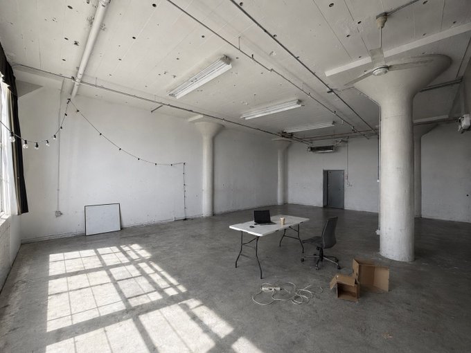 A spacious, empty industrial loft with white walls, concrete floors, large windows casting window pane shadows, featuring a single table with a laptop and chair in the center.