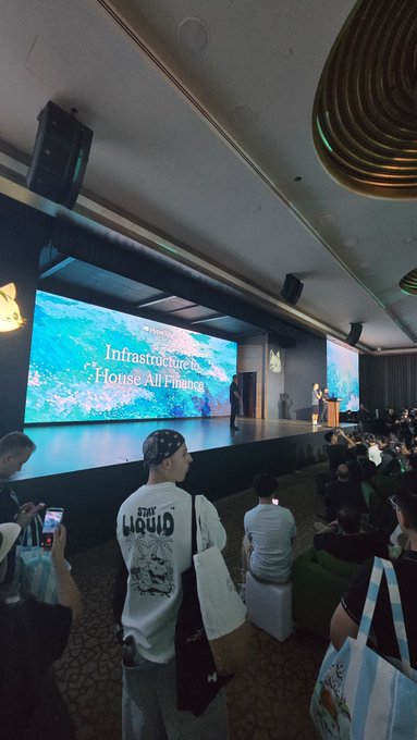 A crowd gathered at an event with a stage screen displaying 'Infrastructure House All Hands,' with attendees including someone wearing a 'STAY LIQUID' shirt.