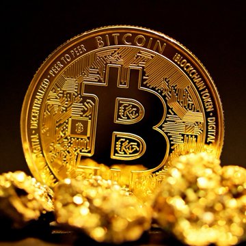 In the image, there are three gold coins displayed together against a dark background. The middle coin features the Bitcoin logo and text that says "BITCOIN". Below this main coin, there is another smaller coin with a similar design. The largest of these smaller coins also has the Bitcoin logo on it. In addition to the coins themselves, there are some small rocks visible in the bottom right corner of the image.