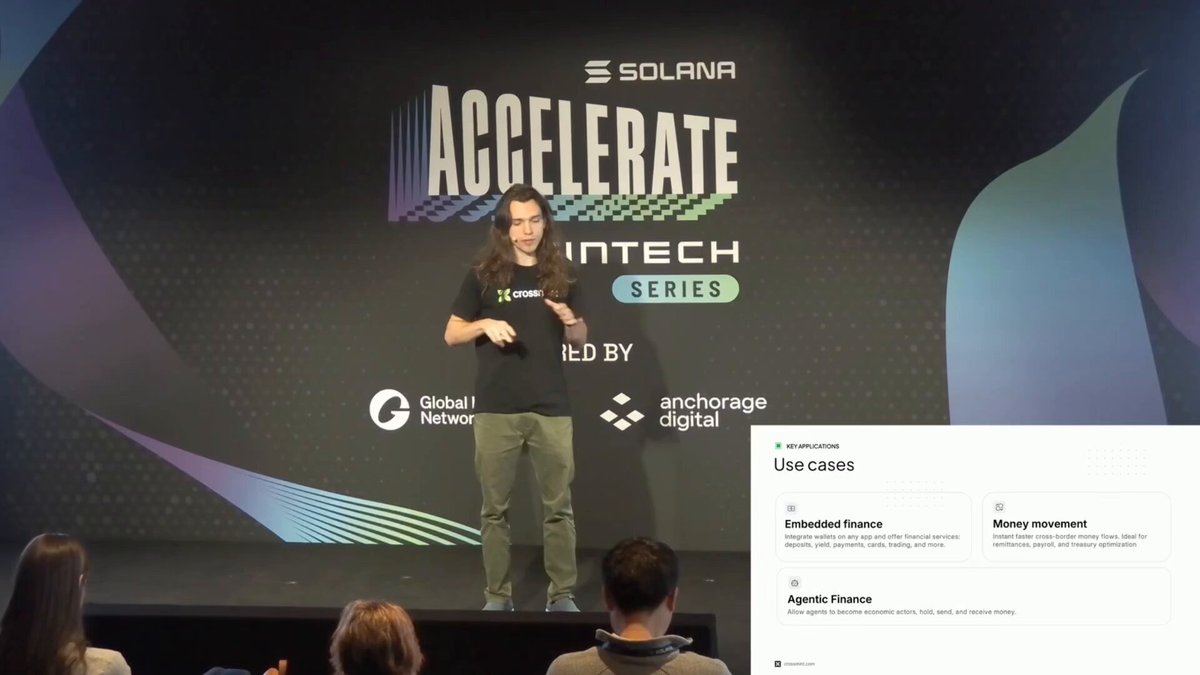 The image shows a man standing at a podium during an event, giving a speech or presentation. The backdrop of the scene features a large screen displaying text related to the Accelerate InTech Series. There are several people present in the background, some sitting on chairs and others standing around the stage area. A person is also seen holding a cell phone near one of the attendees.