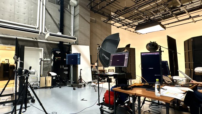 A photograph of a professional photography studio with lighting equipment, softboxes on tripods, computer monitors, backdrops, and exposed ceiling rigging.