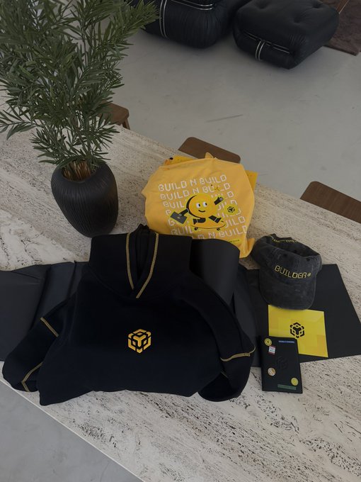 The image features a table with various items, including a black hoodie with a yellow logo, a black backpack, a black vase holding a plant, and some other objects such as a book and a keychain. The background of the image is blurred, but there are chairs visible in the scene. The main focus of the image is on the table and its contents, which include items related to building or construction.