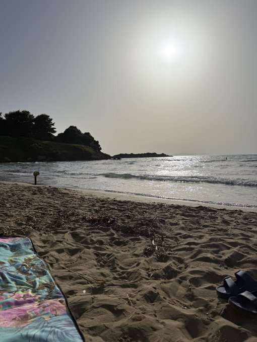 A serene beach photograph at sunset/sunrise, with a colorful patterned towel and sandals in the foreground, gentle waves lapping the sandy shore, and a tree-lined coastline in the distance.