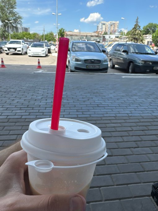 A hand holding a bubble tea cup with a red straw and sealed plastic film lid, with parked cars and a sunny outdoor area visible in the background.