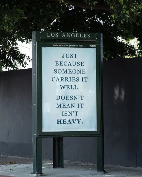 An outdoor Los Angeles sign displaying the motivational quote: 'JUST BECAUSE SOMEONE CARRIES IT WELL, DOESN'T MEAN IT ISN'T HEAVY.' with the tagline 'Make sure your friends are okay.'