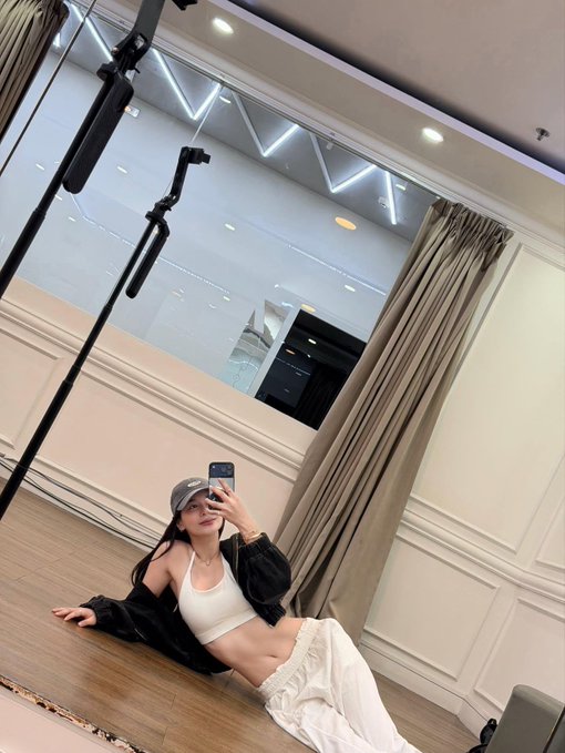 A woman takes a mirror selfie while sitting on the floor of a room, wearing a white sports bra, white pants, an open black shirt, and a cap.