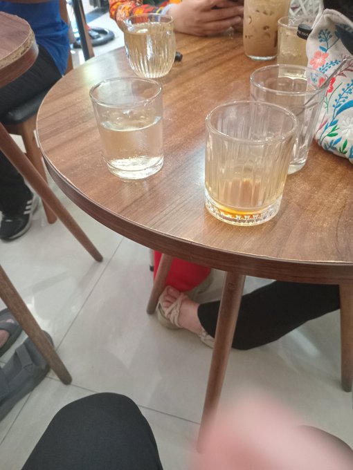 A wooden cafe table holds several glasses of water and iced coffee, with people's legs and sandals visible underneath on a tiled floor.