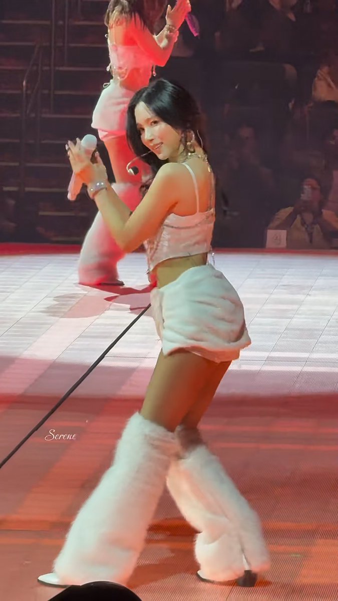 A member of the K-pop group TWICE, specifically MINA, performs on stage in a pink two-piece outfit with fluffy leg warmers, holding a microphone while dancing. The watermark "Serene" is visible in the lower left, and the image captures her mid-performance with red stage lighting illuminating the scene.