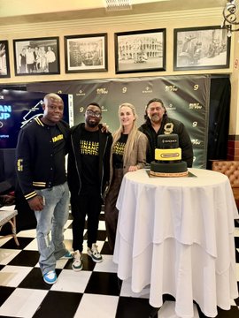 Four people posing together at an event standing next to a celebratory cake on a white-draped table, with framed photos on the wall behind them.