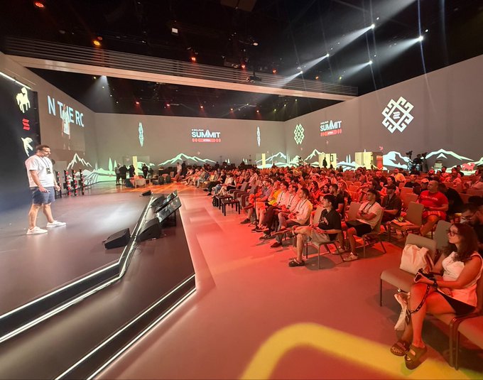 A presenter stands on stage at a cryptocurrency summit in front of a large seated audience, with "SUMMIT" branding visible on the screens behind.