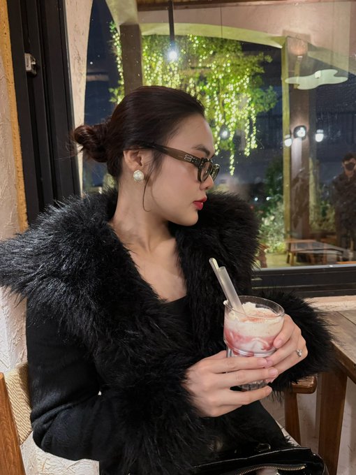 The image features a woman sitting at a table with a drink, wearing glasses and a fur coat. She appears to be enjoying her time as she looks off into the distance. The background of the photo shows a window that provides natural light for the scene. There is also an element of social media presence in the image, as there are several people visible in the background who might have taken the picture or posted it on social media platforms.