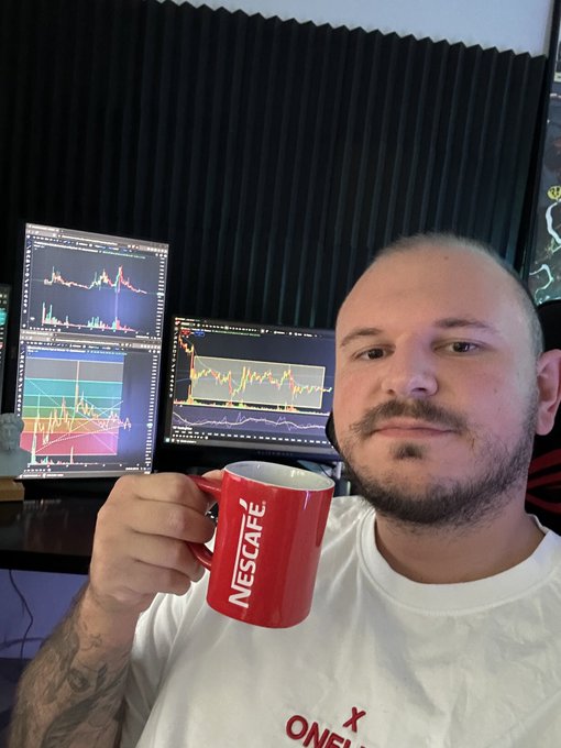 A man holds a red Nescafé mug while sitting in front of multiple computer monitors displaying trading charts and candlestick graphs, suggesting a day trading or financial analysis setting.