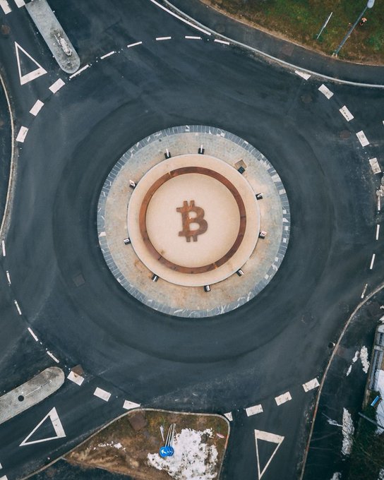 An aerial view of a roundabout in Slovenia featuring a Bitcoin symbol (₿) painted on its center, marking the world's first Bitcoin monument. The dark asphalt encircles the tan-colored monument, with directional arrows and lane markings guiding traffic around this historic crypto landmark.