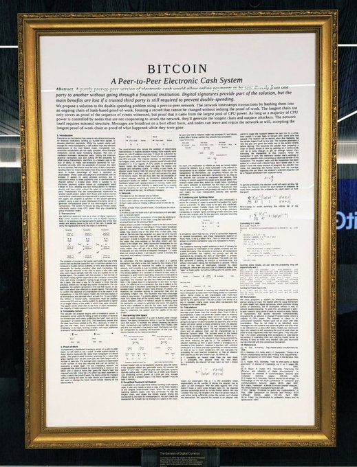 A framed copy of the Bitcoin whitepaper titled 'BITCOIN: A Peer-to-Peer Electronic Cash System' by Satoshi Nakamoto hangs on a wall, displayed in an ornate gold frame.